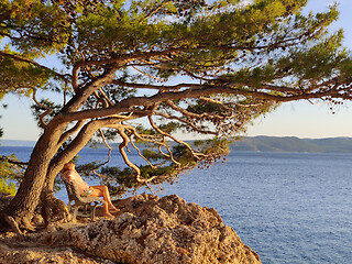 Image showing Pensive woman on vacations, sitting and relaxing under large pine tree on bench by dip blue sea enjoying beautiful sunset light in Brela, Makarska region, Dalmatia, Croatia