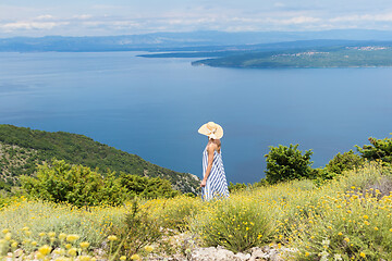 Image showing Rear view of young woman wearing striped summer dress and straw hat standing in super bloom of wildflowers, relaxing while enjoing beautiful view of Adriatic sea nature, Croatia