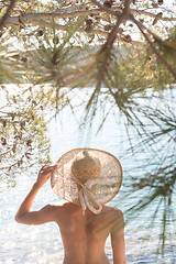 Image showing Rear view of topless beautiful woman wearing nothing but straw sun hat realaxing on wild coast of Adriatic sea on a beach in shade of pine tree.