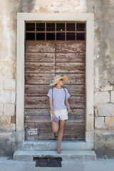 Image showing Beautiful young female tourist woman standing in front of vinatage wooden door and textured stone wall at old Mediterranean town, smiling, holding, smart phone to network on vacationes