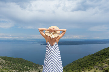 Image showing Rear view of young woman wearing striped summer dress and straw hat standing in super bloom of wildflowers, relaxing while enjoing beautiful view of Adriatic sea nature, Croatia