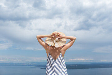 Image showing Rear view of young woman wearing striped summer dress and straw hat standing in super bloom of wildflowers, relaxing while enjoing beautiful view of Adriatic sea nature, Croatia