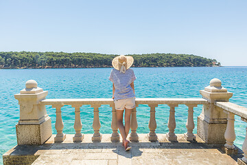 Image showing Rear view of woman wearing straw summer hat ,leaning against elegant old stone fence of coastal villa, relaxing while looking at blue Adriatic sea, on Losinj island Croatia.