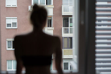 Image showing Home quarantine and social distancing during covid pandemic. Silhouette of lonley caucasian woman standing by window, anxiously looking out. Coronavirus infection, pandemics, disease outbreaks