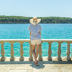 Image showing Rear view of woman wearing straw summer hat ,leaning against elegant old stone fence of coastal villa, relaxing while looking at blue Adriatic sea, on Losinj island Croatia.