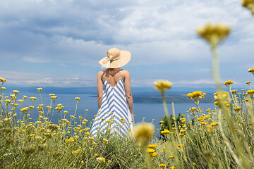 Image showing Rear view of young woman wearing striped summer dress and straw hat standing in super bloom of wildflowers, relaxing while enjoing beautiful view of Adriatic sea nature, Croatia