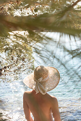 Image showing Rear view of topless beautiful woman wearing nothing but straw sun hat realaxing on wild coast of Adriatic sea on a beach in shade of pine tree.