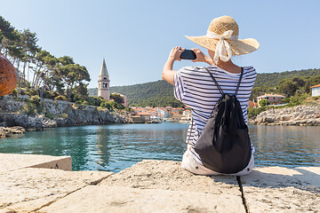 Image showing Woman traveler wearing straw summer hat and backpack, sittingat edge of stone pier, taking photo of beautiful panoramic view of Veli Losinj, Losinj island, Croatia