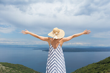 Image showing Rear view of young woman wearing striped summer dress and straw hat standing in super bloom of wildflowers, relaxing with hands up to the sky, enjoing beautiful view of Adriatic sea nature, Croatia