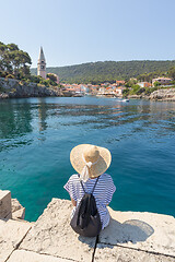 Image showing Woman traveler wearing straw summer hat and backpack, sittingat edge of stone pier, enjoying beautiful panoramic view of Veli Losinj, Losinj island, Croatia.