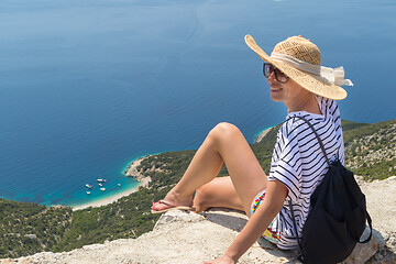 Image showing Active sporty woman on summer vacations sitting on old stone wall at Lubenice village, wearing straw hat and beach backpack enjoying beautiful coastal view of Cres island, Croatia