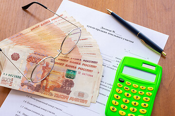 Image showing On the table is a contract for the sale of a land plot, a bundle of money, a calculator, a pen and glasses