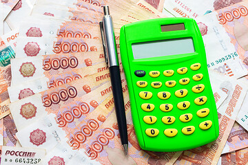 Image showing A green calculator and a ballpoint pen lie on a fanned stack of five thousandth bills