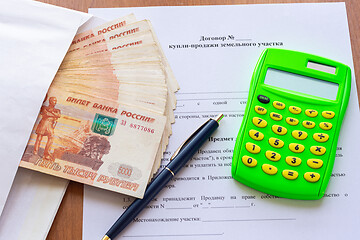 Image showing On the table is a contract for the sale of a land plot, a bundle of money in an envelope, a pen and a calculator