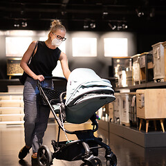 Image showing Young mom with newborn in stroller shopping at retail furniture and home accessories store wearing protective medical face mask to prevent spreading of corona virus. New normal during covid epidemic