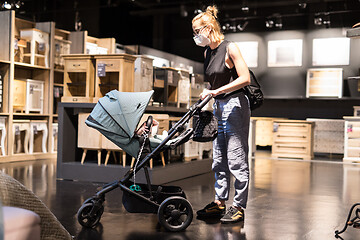 Image showing Young mom with newborn in stroller shopping at retail furniture and home accessories store wearing protective medical face mask to prevent spreading of corona virus. New normal during covid epidemic