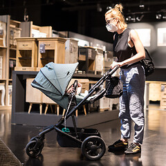 Image showing Young mom with newborn in stroller shopping at retail furniture and home accessories store wearing protective medical face mask to prevent spreading of corona virus. New normal during covid epidemic