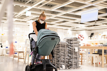 Image showing Young mom with newborn in stroller shopping at retail furniture and home accessories store wearing protective medical face mask to prevent spreading of corona virus. New normal during covid epidemic