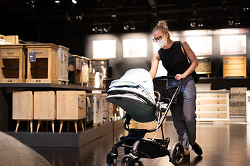 Image showing Young mom with newborn in stroller shopping at retail furniture and home accessories store wearing protective medical face mask to prevent spreading of corona virus. New normal during covid epidemic