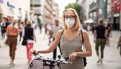 Image showing Woman walking by her bicycle on pedestrian city street wearing medical face mask in public to prevent spreading of corona virus. New normal during covid epidemic.