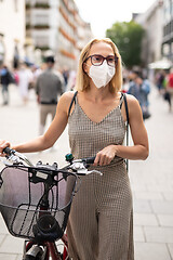 Image showing Woman walking by her bicycle on pedestrian city street wearing medical face mask in public to prevent spreading of corona virus. New normal during covid epidemic.