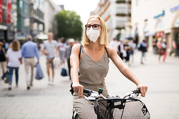 Image showing Woman riding bicycle on city street wearing medical face mask in public to prevent spreading of corona virus. New normal during covid epidemic.