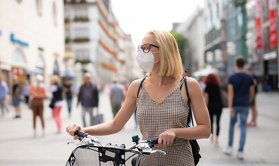 Image showing Woman walking by her bicycle on pedestrian city street wearing medical face mask in public to prevent spreading of corona virus. New normal during covid epidemic.