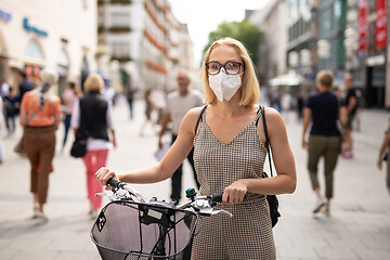 Image showing Woman walking by her bicycle on pedestrian city street wearing medical face mask in public to prevent spreading of corona virus. New normal during covid epidemic.