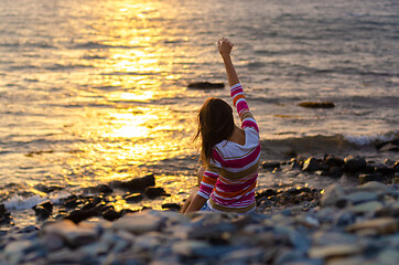 Image showing A girl sits on the seashore and throws stones into the water, raised her hands, evening, sunset