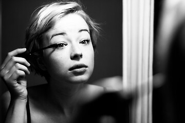 Image showing Woman getting ready for work doing morning makeup routine applying mascara in bathroom mirror at home. Beautiful caucasian girl applying eye make-up. Black and white image