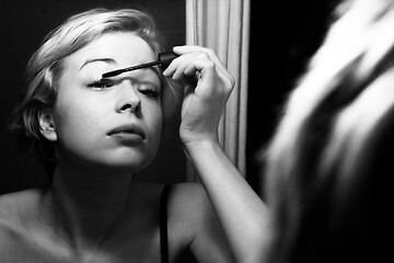 Image showing Woman getting ready for work doing morning makeup routine applying mascara in bathroom mirror at home. Beautiful caucasian girl applying eye make-up. Black and white image