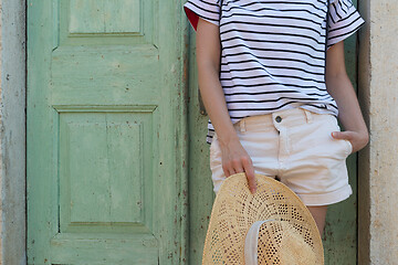 Image showing Fashinable torso portrait of woman holding sun hat, standing and relaxing in shade in front of turquoise vinatage wooden door in old Mediterranean town while sightseeing on hot summer day