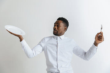 Image showing Young doubting attractive african american guy holding empty dish and fork isolated on grey background. Copy space and mock up. Blank template background.