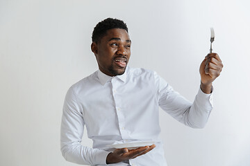 Image showing Young african american guy holding empty dish and fork with disgusting facial expression isolated on grey background. Copy space and mock up. Blank template background.