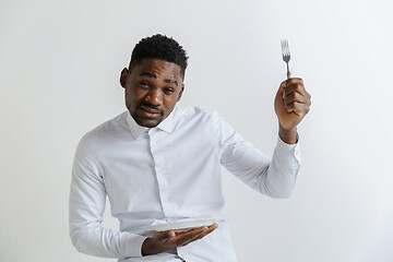 Image showing Young doubting attractive african american guy holding empty dish and fork isolated on grey background. Copy space and mock up. Blank template background.