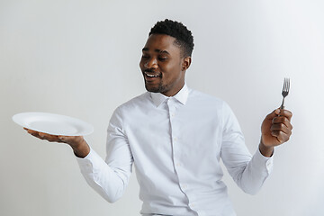 Image showing Young doubting attractive african american guy holding empty dish and fork isolated on grey background. Copy space and mock up. Blank template background.