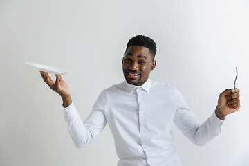 Image showing Young doubting attractive african american guy holding empty dish and fork isolated on grey background. Copy space and mock up. Blank template background.