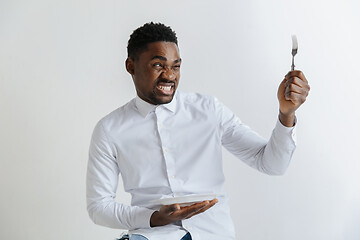 Image showing Young african american guy holding empty dish and fork with disgusting facial expression isolated on grey background. Copy space and mock up. Blank template background.