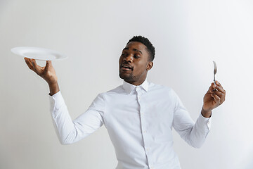 Image showing Young doubting attractive african american guy holding empty dish and fork isolated on grey background. Copy space and mock up. Blank template background.