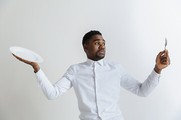 Image showing Young doubting attractive african american guy holding empty dish and fork isolated on grey background. Copy space and mock up. Blank template background.