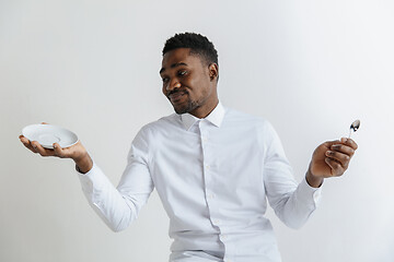 Image showing Young doubting attractive african american guy holding empty dish and spoon isolated on grey background. Copy space and mock up. Blank template background.