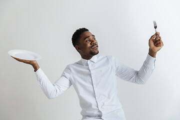 Image showing Young doubting attractive african american guy holding empty dish and fork isolated on grey background. Copy space and mock up. Blank template background.