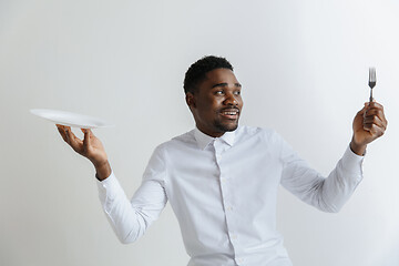 Image showing Young doubting attractive african american guy holding empty dish and fork isolated on grey background. Copy space and mock up. Blank template background.