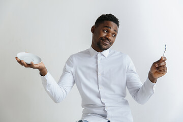 Image showing Young doubting attractive african american guy holding empty dish and spoon isolated on grey background. Copy space and mock up. Blank template background.