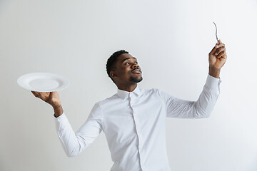 Image showing Young doubting attractive african american guy holding empty dish and fork isolated on grey background. Copy space and mock up. Blank template background.