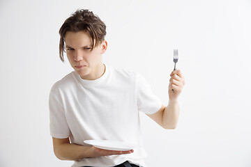 Image showing Young sad attractive guy holding empty dish and fork isolated on grey background.