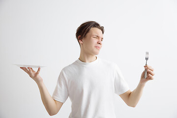 Image showing Young sad attractive guy holding empty dish and fork isolated on grey background.