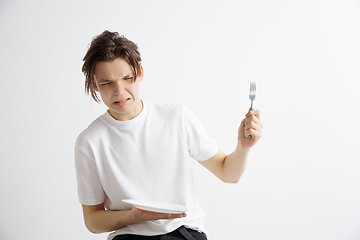 Image showing Young sad attractive guy holding empty dish and fork isolated on grey background.