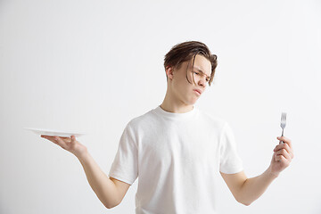 Image showing Young sad attractive guy holding empty dish and fork isolated on grey background.