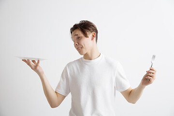 Image showing Young sad attractive guy holding empty dish and fork isolated on grey background.